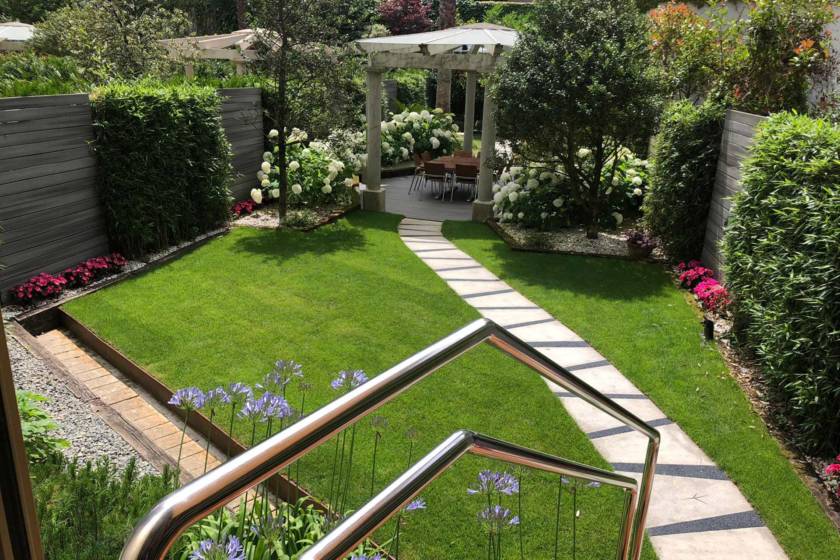 jardín con el césped recién cortado, jardín decorado con un paseo hecho de piedra, hortensias blancas y flores de color morado y rosa que llevan a una mesa de madera tapada por unos pilares